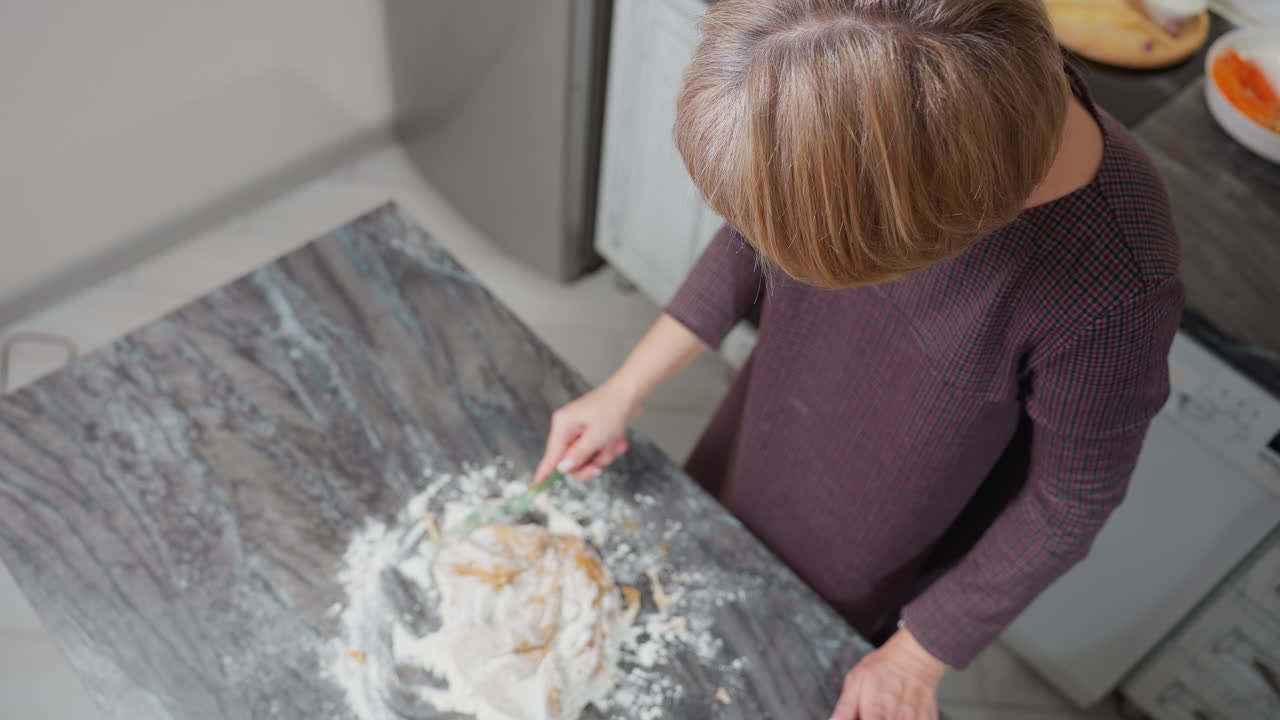 Aerial view of person kneading dough on floured tiled table in kitchen, hands work on soft, sticky dough as flour spreads across countertop, traditional homemade baking process