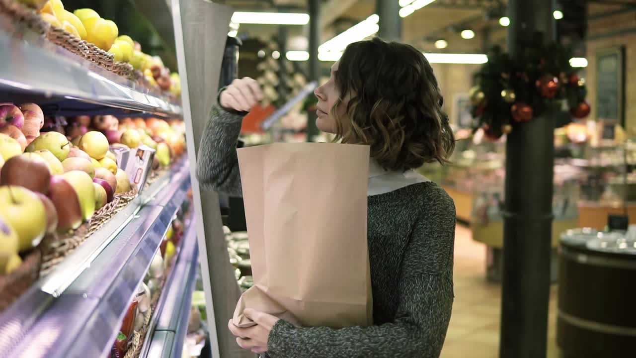 una niña linda compra manzanas rojas frescas en el mercado. una hermosa mujer joven se para frente al estante y pone las manzanas en un marrón