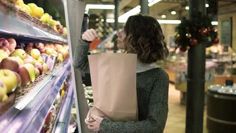 Cute-girl-buys-fresh-red-apples-in-the-market.-Beautiful-young-woman-stands-in-front-the-shelf-and-puts-the-apples-to-a-brown