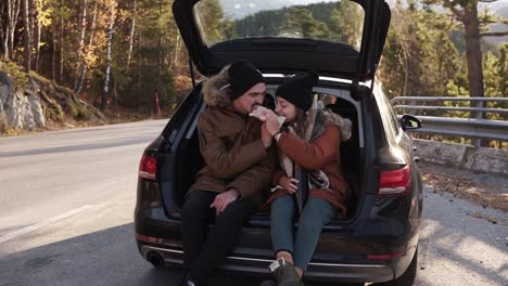 una pareja de enamorados en un viaje por carretera. el chico y la chica están sentados en el maletero abierto del coche, comiendo sándwiches y alimentándose mutuamente. detenerse al aire libre en medio de la carretera. día frío y soleado de otoño