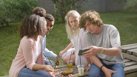 Young-Man-Showing-Something-Interesting-On-Mobile-Phone-To-His-Multiethnic-Friends-In-A-Park