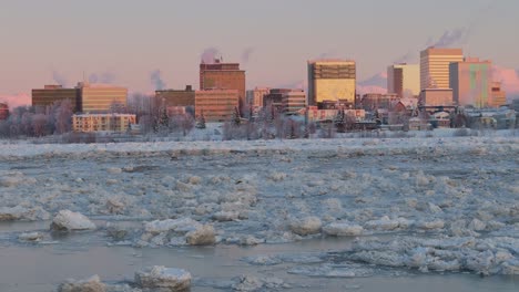 slowly drone dolley shot over a frozen lake with in the background the skyline of anchorage illuminated by sunset