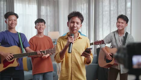 young asian boy, in front of his friends with musical instruments, holding a microphone and reading comments on smartphone while live stream at home