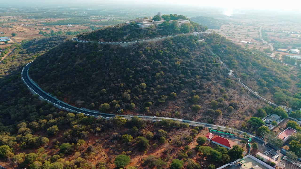 Cinematic aerial view of Sivanmalai Temple majestically perched atop a hill, surrounded by greenery, with a winding mountain road adding charm to this serene spiritual and scenic landmark