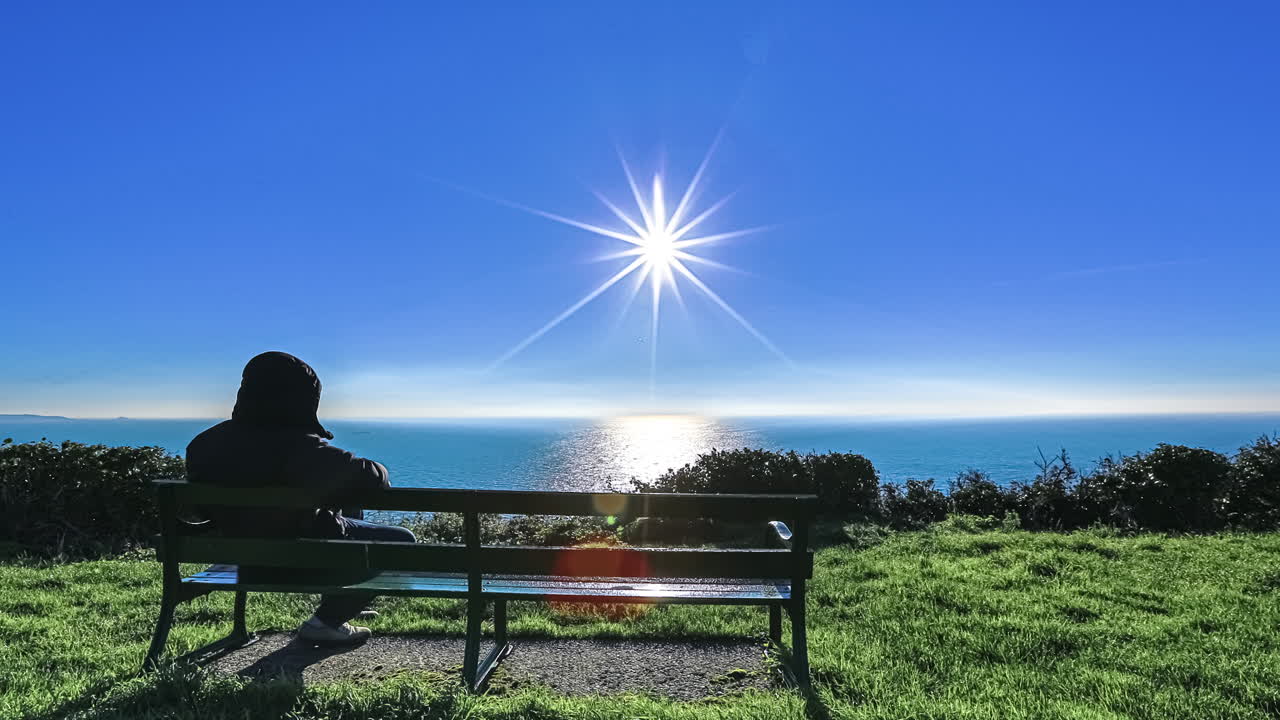 Time lapse shot of man sitting on bench and enjoying sunrise over blue ocean and blue sky in summer