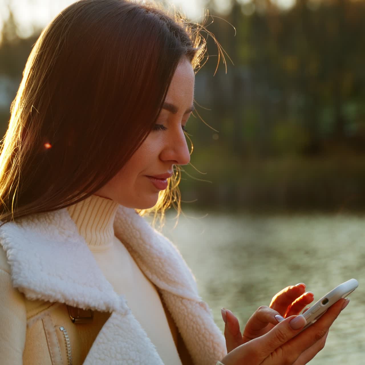 Beautiful dark-haired lady typing on her phone. Relaxed Caucasian woman using her phone standing at the river bank on warm autumn day