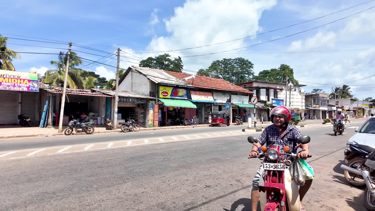 A traditional red bus travels through a local town in Sri Lanka, with a motorcyclist riding in the foreground. The image captures daily life and transportation.