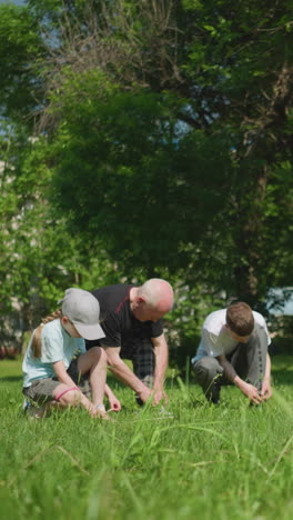 grandparents and their grandsons are all kneeling on a grassy field, tying their shoelaces, the youngest boy is resting his head on a soccer ball, with his brother in red looking at him