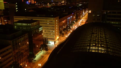 a high-angle timelapse unveils a night cityscape, foregrounded by a large, lit glass-roofed building, set against a backdrop of glowing street-lit streets and illuminated buildings