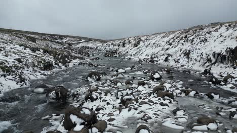 Ein-Fluss-Am-Straßenrand-Mit-Schneebedeckten-Felsen-Und-Kaskadierendem-Wasser-Im-Norden-Islands-Im-Winter,-Aus-Der-Luft