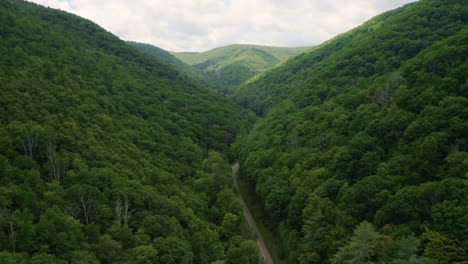 Aerial-drone-flying-forward-through-green-summer-forest-as-rural-mountain-road-carves-through-the-valley-with-blue-skies-and-clouds-overhead