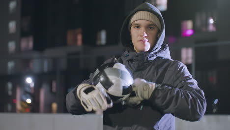 coach rolling soccer ball in his hands while wearing hoodie and gloves at an urban sports field during nighttime practice, dimly lit buildings and bokeh lights