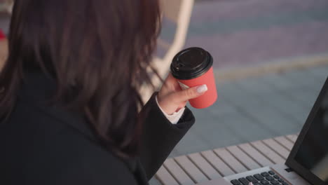 primer plano de un estudiante con abrigo negro bebiendo una taza de café roja al aire libre mientras escribe en una computadora portátil, colocando la taza hacia abajo con enfoque en el trabajo, el fondo presenta una silla y un entorno urbano