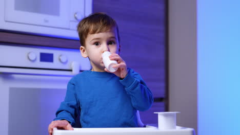 caucasian toddler wearing blue sweater drinks from a little white bottle. baby boy drinking dairy in the kitchen.