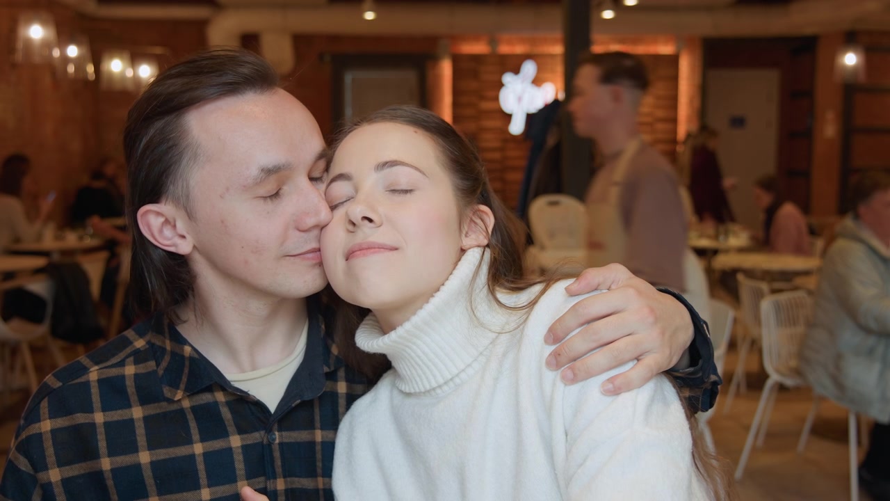 Close-up de homem e mulher posando juntos em iluminação suave, com a câmera capturando suas expressões enquanto estão próximos um do outro. O fundo mostra um ambiente interno com pessoas jantando.