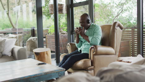 happy african american senior man relaxing in armchair, wearing headphones and drinking coffee