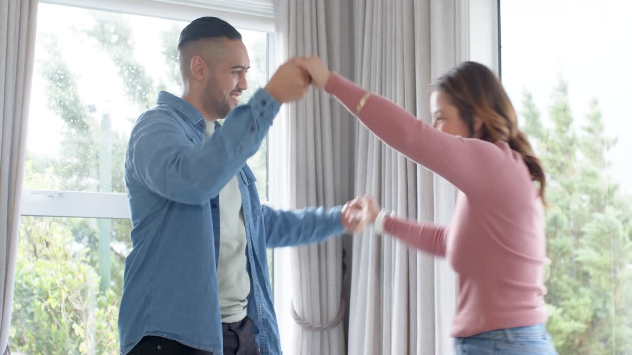 Diverse couple rising from wooden table pasta breakfast as man's words prompt dancing by wet window