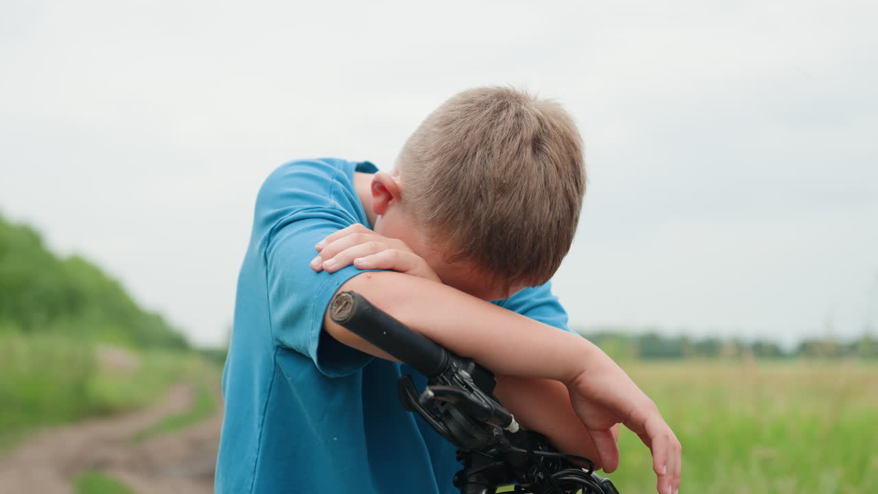 Boy rests quietly beside bike outdoors, Teenager stands pensively with bicycle beneath cloudy sky in nature, Young boy leaning on his bicycle amidst lush green surroundings on cloudy summer day