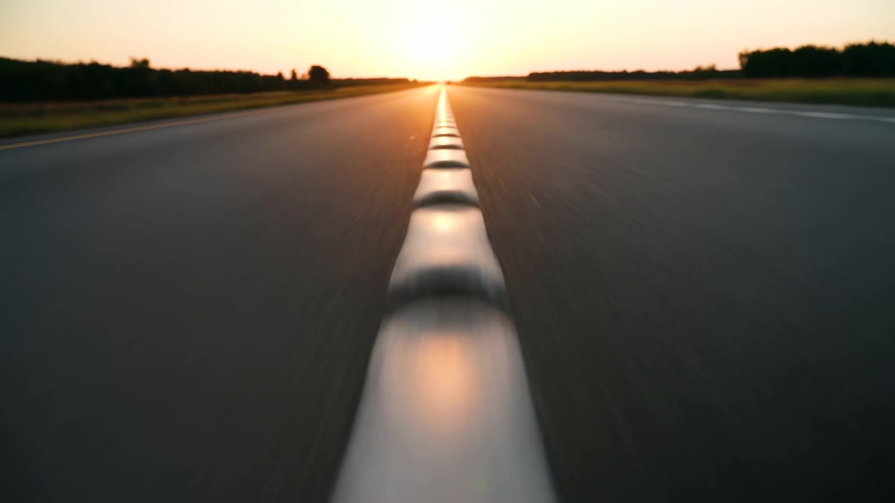 Fremoverbevegelse følger hvite kjørefeltmarkeringer på en rolig motorvei ved soloppgang, med glødende horisont, mørke silhuetter langs veien og en sentrert komposisjon som fremhever dybde, avstand og glatt veibane