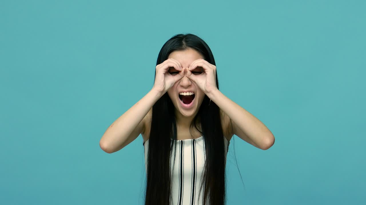 긴 검은 머리카락을 가진 흥미진진한 아시아 여성은 안경으로 손가락을 통해 쳐다보고 있습니다.