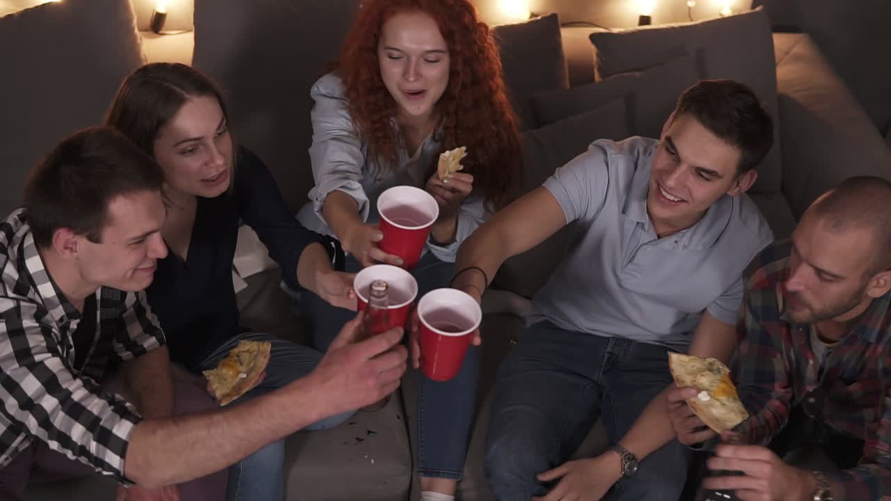 Happy friends are celebrating new years eve party at home. Pizza, bottles of beer and red cups - clinking, cheers. Holidays and celebration concept. High angle view
