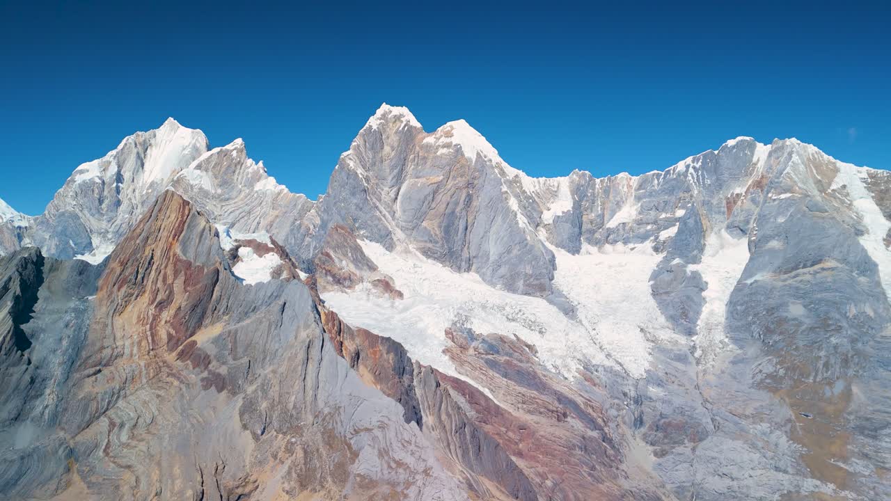Drone shot flying over jagged snow-covered peaks and colorful rocky slopes of the Cordillera Huayhuash under a clear blue sky