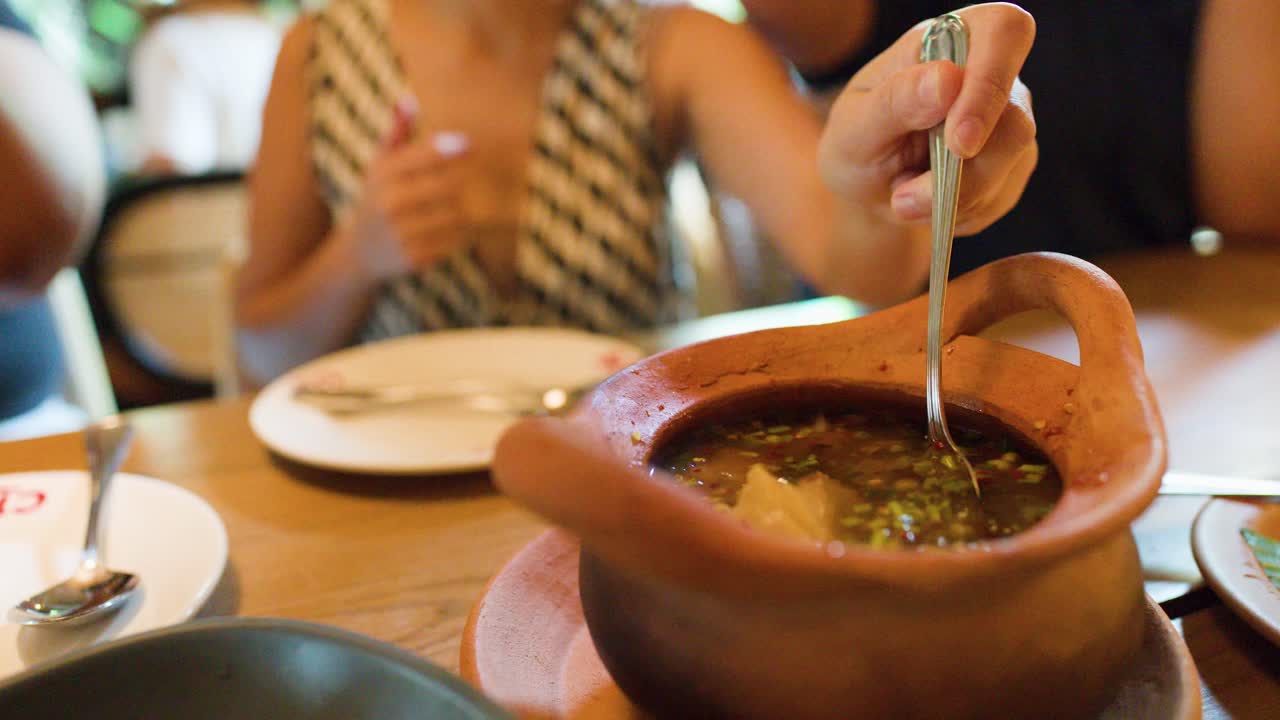 Woman ladles herbal Thai soup from clay pot in warmly lit Bangkok dining setting