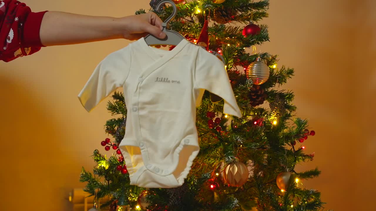 Baby announcement showing a bodysuit with "Little One" written on it in front of the illuminated Christmas tree