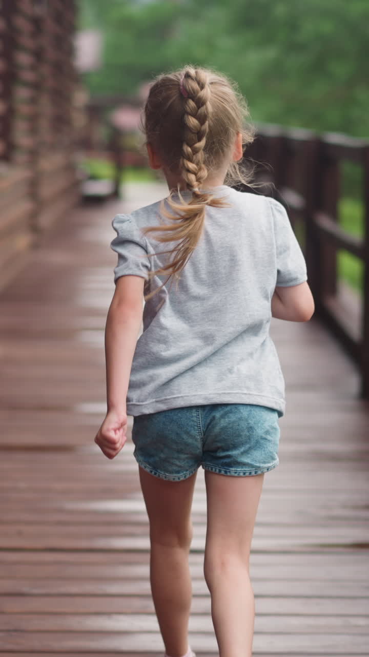 Little girl with blonde plait runs playing along empty veranda deck at suburban cottage house on spring day backside view slow motion