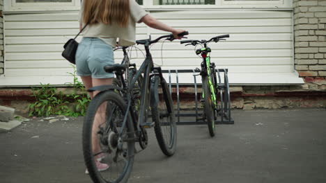 back view of young woman in pink sneakers, jean shorts, and black handbag moving bicycle toward bicycle stand, setting bike among other parked bicycles with white building backdrop