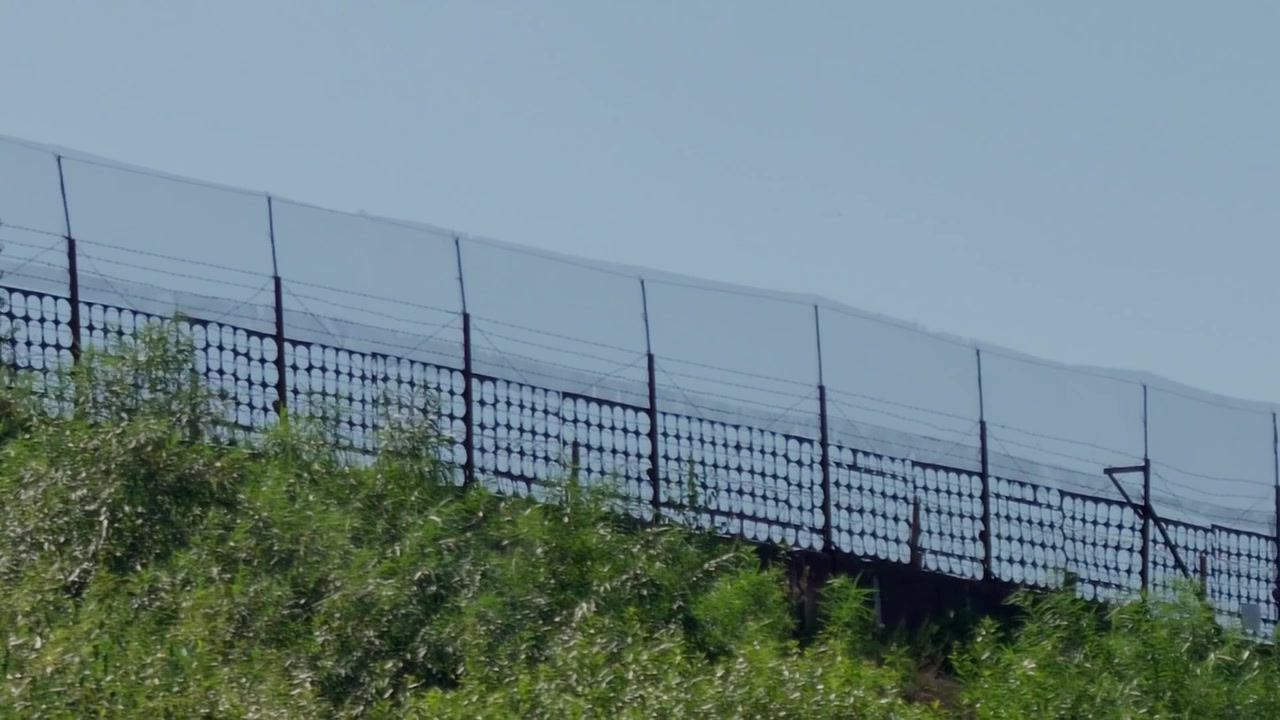 Una ripresa in movimento cattura una recinzione di confine ad alta sicurezza con filo spinato e rete metallica che si erge su una collina erbosa contro un cielo sereno in Lettonia, evidenziando la protezione dei confini