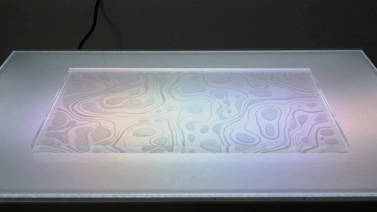Lightbox alimentato che illumina linee incise su un pannello satinato, con colori cangianti in uno studio in penombra, cavo visibile.