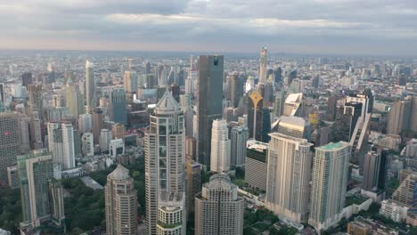 Aerial-through-skyline-and-skyscrapers-of-Sukhumvit-district,-Pathum-Wan-and-Lumphini-Park-during-sunset-in-Bangkok,-Thailand