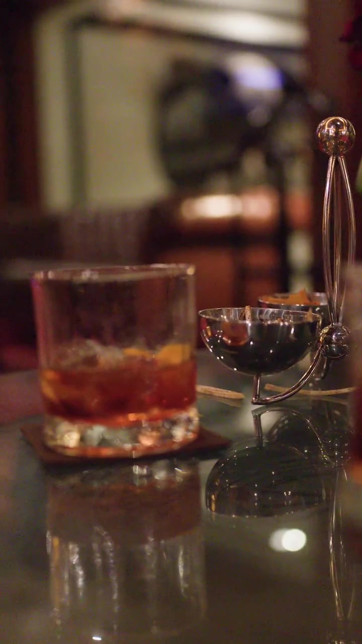 Bicchiere di whisky con ghiaccio su tavolo da bar riflettente, illuminazione ambientale calda, messa a fuoco ridotta, ambiente elegante
