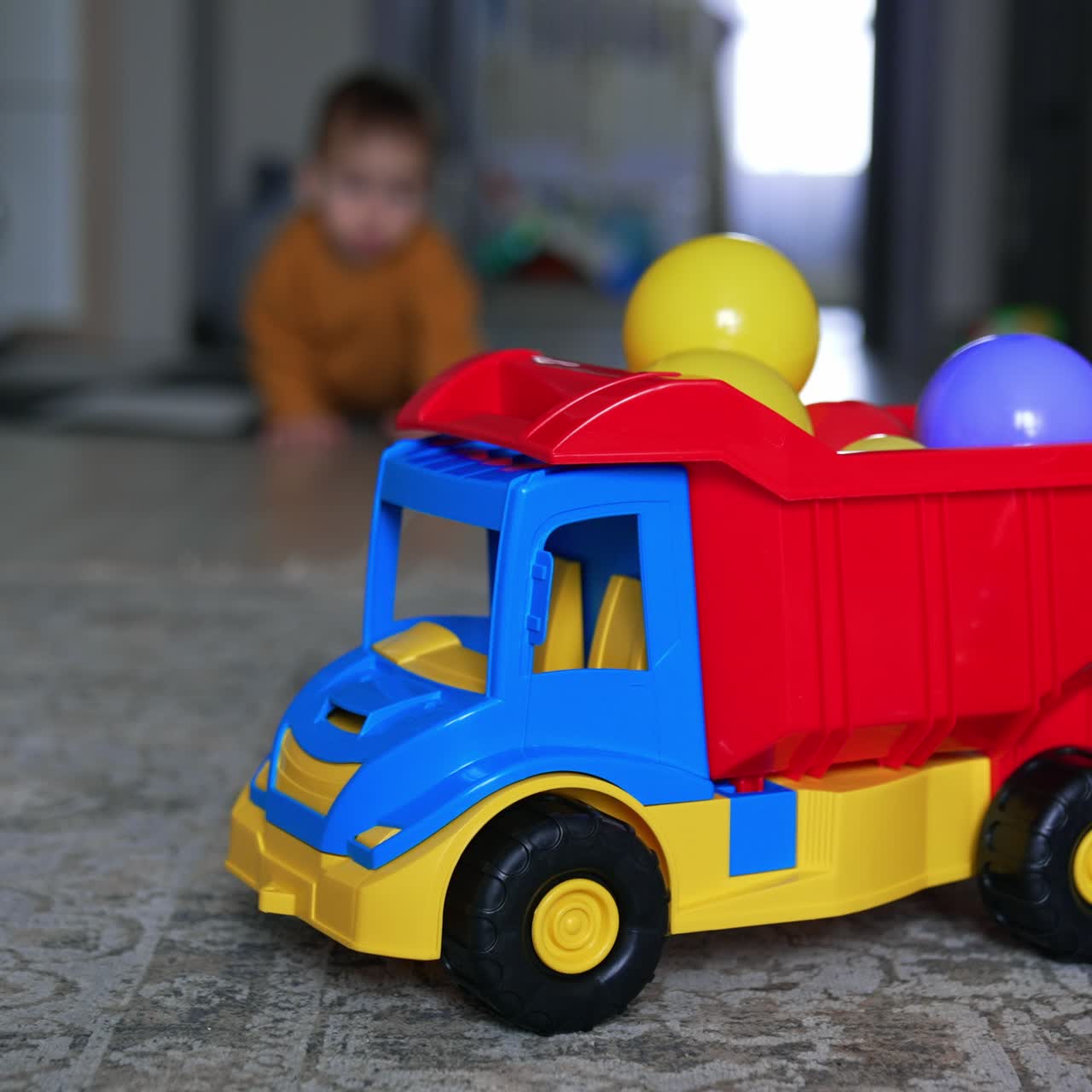 Colorful toy car with balls on the floor in the living room. Little baby appears in the room and crawls to the foreground. Blurred backdrop