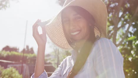 video portrait of happy biracial woman wearing sunhat in sunny garden smiling to camera