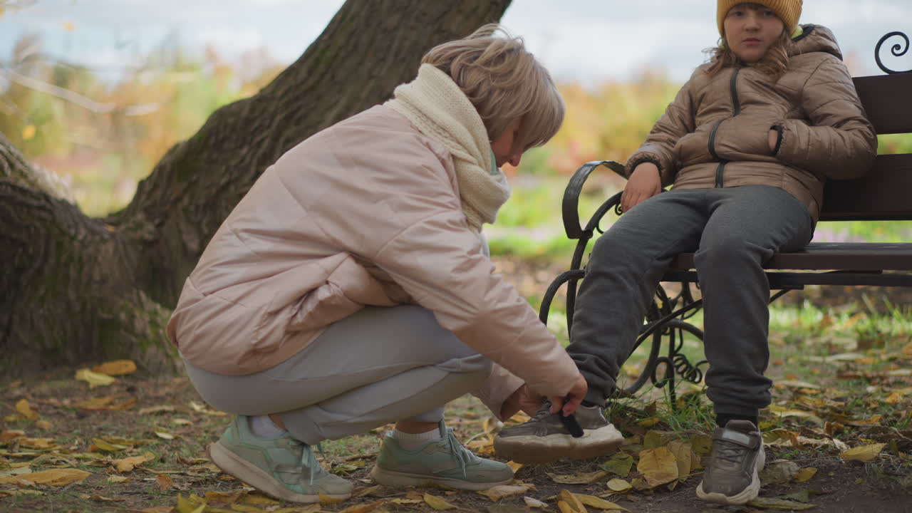 caring mother kneels on fallen leaves fastening kid shoe strap near wooden bench in tranquil autumn park, surrounded by vivid foliage, warm jackets, and peaceful family bonding in outdoor setting