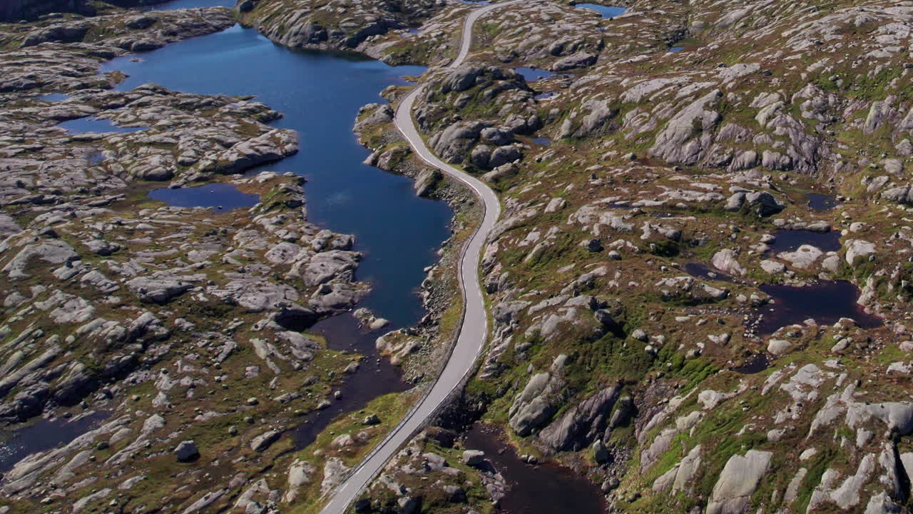 Aerial shot, panning across a winding mountain road as it snakes through the mossy, rocky, lake covered landscape of Rogaland in Norway
