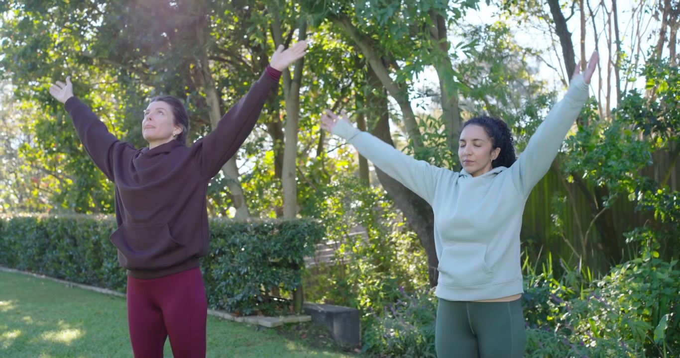 Wanita dari berbagai latar belakang menarik napas sambil menyatukan telapak tangan dalam posisi berdoa, berlatih yoga mengenakan hoodie dan legging di halaman belakang.