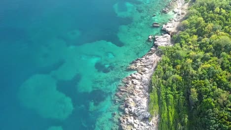 Fliegen-über-Die-Insel-Ksamil-Mit-Den-Bäumen,-Die-Auf-Den-Felsigen-Inselrand-Und-Das-Schöne-Aquarell-Der-Adria-Treffen