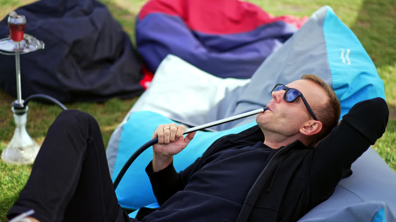 Relaxed man in black clothes and sunglasses lies in bean bag chair outdoors. Resting man smoking hookah producing smoke.