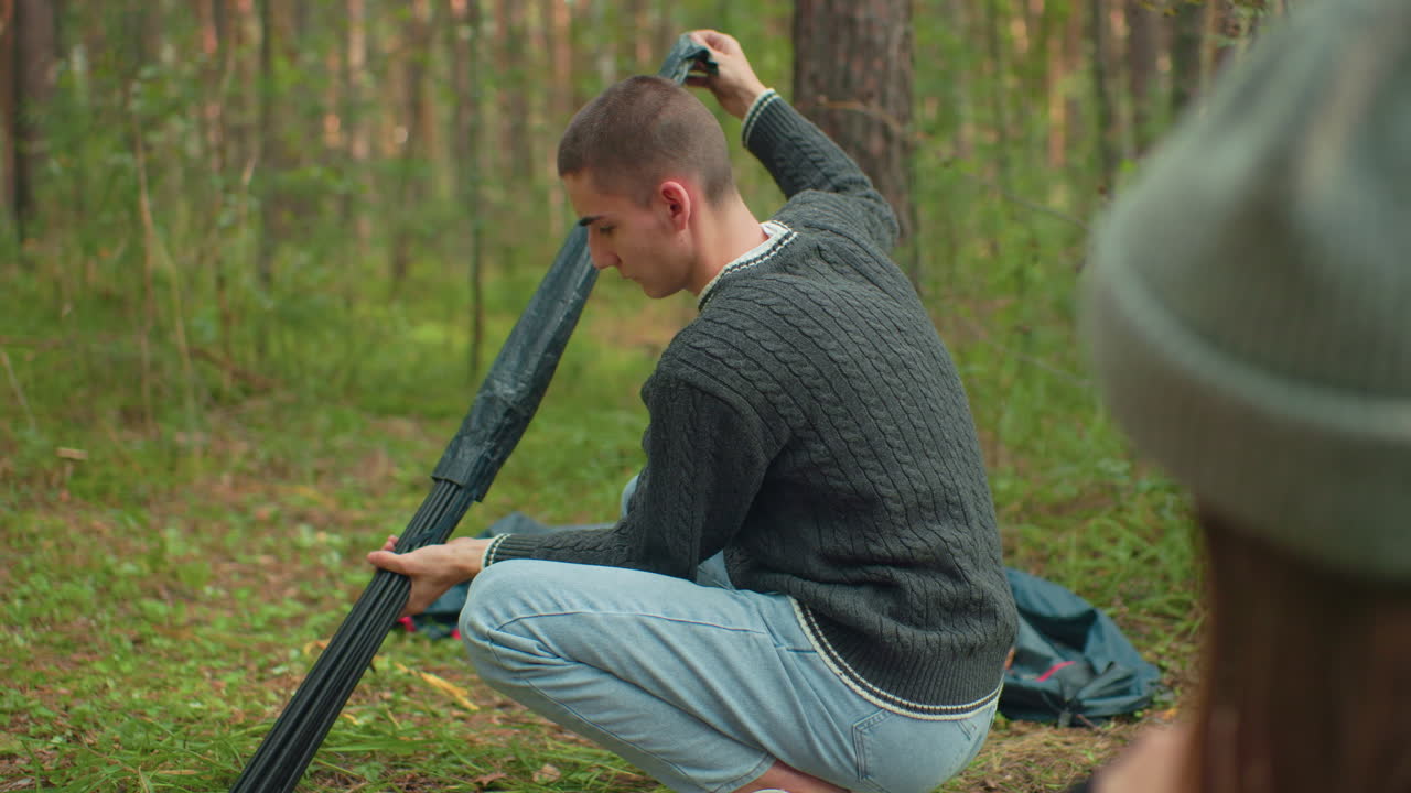 Close up of woman in beanie watching man carefully pull tent pole from black bag while setting up camp in forest, surrounded by tall trees and green grass during peaceful outdoor moment