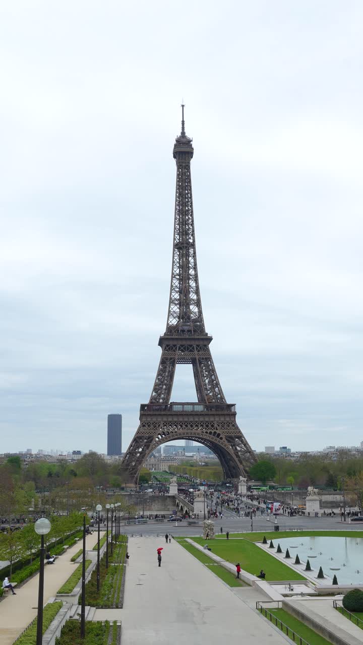 Vista verticale della Torre Eiffel a Parigi con persone che camminano e traffico in movimento sotto. Ripresa diurna che mostra l'area del Campo di Marte e l'attività cittadina.