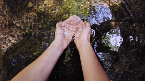 female-hands-holding-water-at-waterfall-stream,-slowmotion,-pov