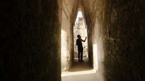 a man in silhouette walks through a narrow stone corridor of a mayan structure in tikal, framed by aged limestone and filtered light.