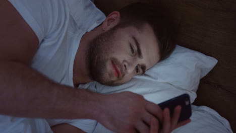 vista superior del hombre caucásico sonriente acostado en la cama, charlando en el teléfono inteligente a altas horas de la noche