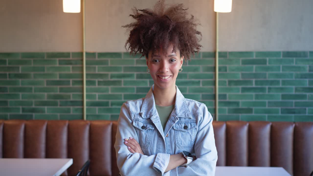 Mujer afroamericana reaccionando al zoom de la cámara, ajustándose la chaqueta vaquera y riendo en una cafetería