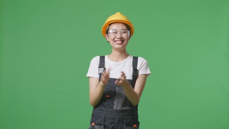 asian woman worker wearing goggles and safety helmet smiling and clapping her hands while standing in the green screen background studio