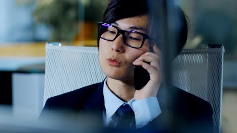 portrait of the japanese businessman wearing suit and glasses, sitting at his desk, talking on mobile phone and working on personal computer.