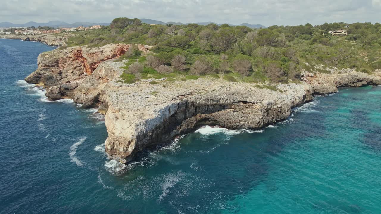 Fly around a dramatic rocky cliff juts into the turquoise sea, surrounded by forest and rugged coastline. The aerial view captures the contrast between the deep blue ocean and the vibrant greenery.